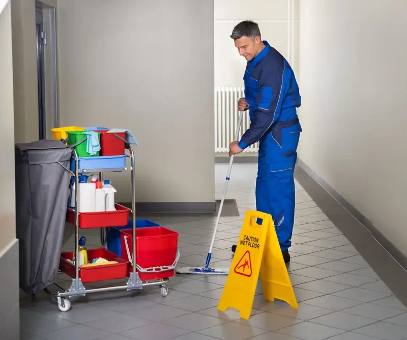 Reinigungskraft beim Wischen eines Flurs mit Putzwagen und Warnschild